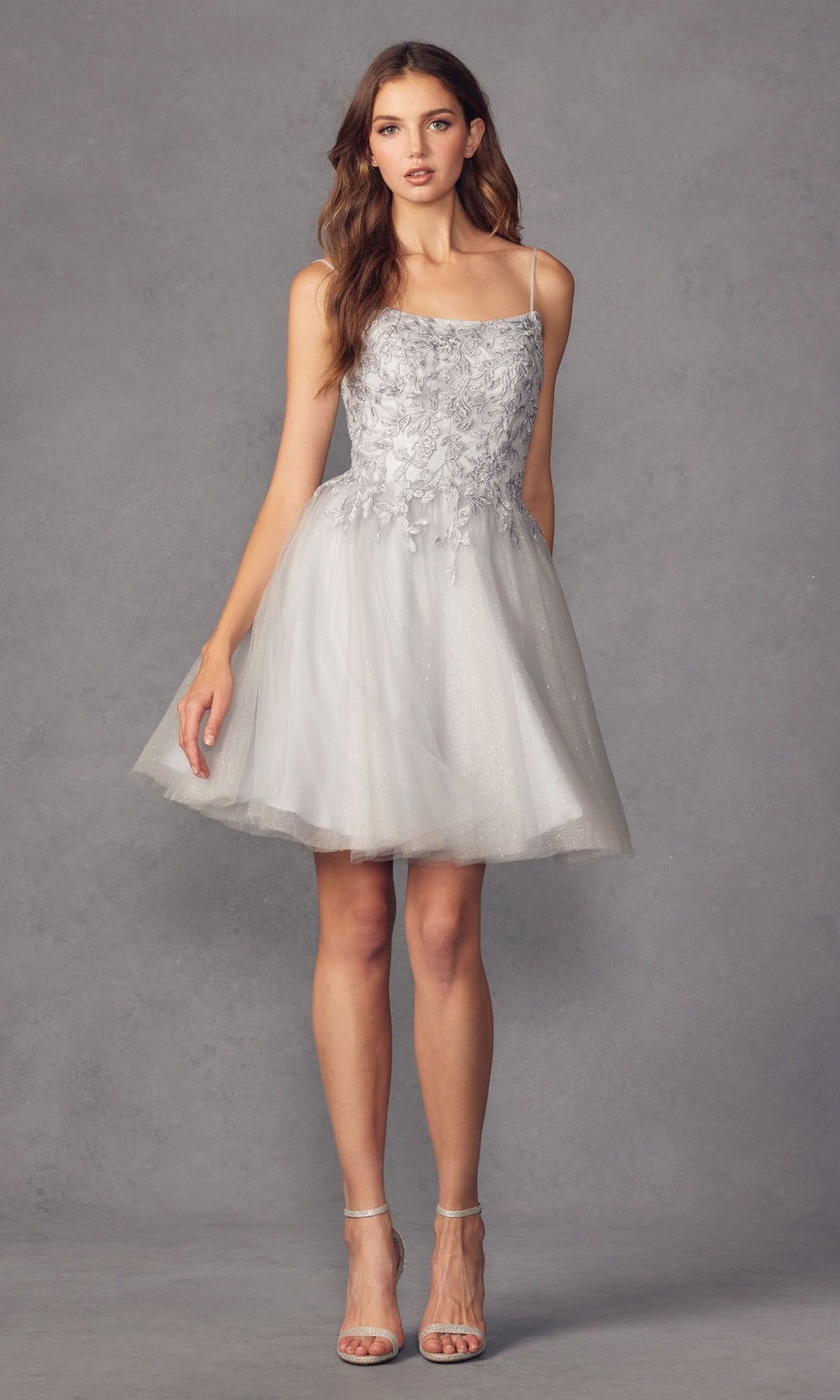 A woman poses against a gray background in the Embroidered-Corset Babydoll Homecoming Dress 860, featuring floral embroidery and thin straps, styled with strappy high-heeled sandals.